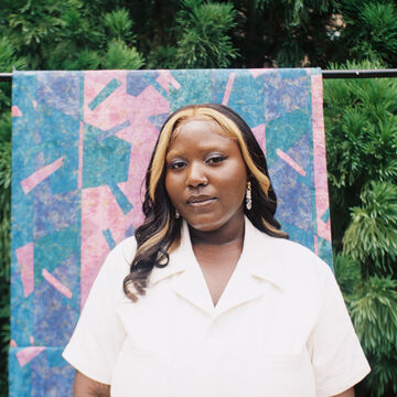 A person with dark skin tone, dark hair with highlights, and wearing a white shirt stands in front of a colorful backdrop surrounded by trees.