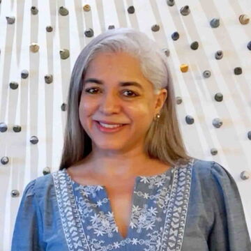 Visiting practitioners Madhvi Subrahmanian, a person with medium skin tone and gray hair, standing against a textured background.