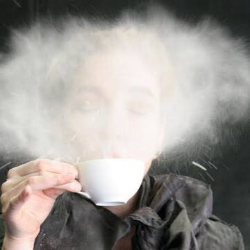 Erica Mott, an adult person with a fair skintone, blowing a substance out of a teacup. 