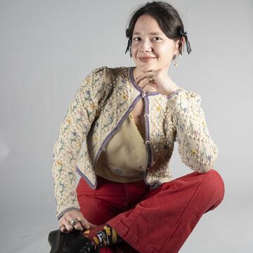 Isabelle Rust sits cross-legged on a stool, smiling in a floral cardigan and red pants.