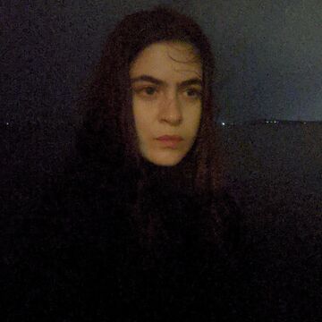Portrait of Azra Keskin, an adult person with a medium-fair skintone, standing outdoors in a dark environment. 