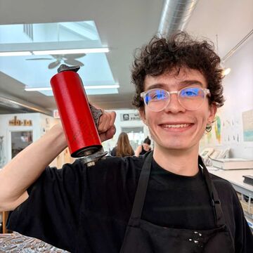 David Lozano smiles while holding a red printing roller in a printmaking studio.