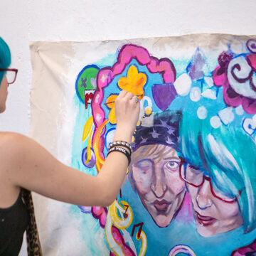 A person with a fair skintone and blue hair paints a colorful artwork of two faces in a heart of symbols. 