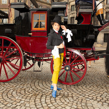 Tommy Walton, an adult person with a fair skintone, tipping their hat in front of a horse and carriage. 