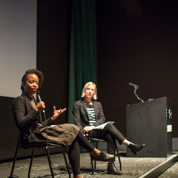 An adult Black person with a medium-deep skintone speaks into a microphone onstage. A person with a fair skintone sits onstage beside them. 