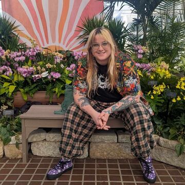 Portrait of MJ Damsen, an adult person with a fair skintone, seated and surrounded by plants. 