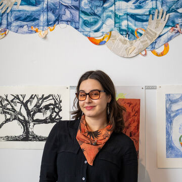 Mya Drost, an adult person with a medium-fair skintone and glasses, standing in front of hanging artwork. 