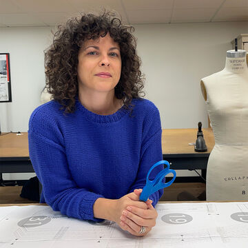 Jonnie Rettele, an adult person with a fair skintone and curly hair, holding a blue pair of scissors that match her sweater. 