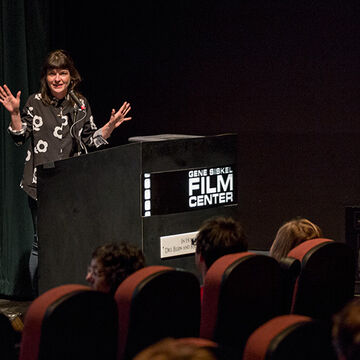 The artist Jodie Mack in a graphic black and white shirt standing with arms raised in front of a podium.