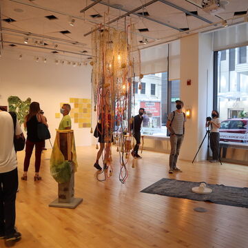 Several people are walking around a room with big windows looking at the artwork installed. Art pieces are on the floor, the wall, and the ceiling.