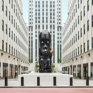 A large black statue of a seated figure with an outsized head in pictured in Rockefeller Plaza