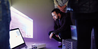 A person crouching down to look at a monitor on the floor. 