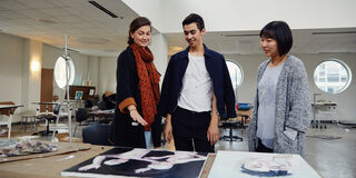 Three people in a large studio room looking at some canvases on a table. 