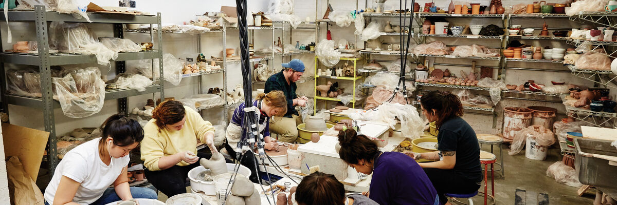 A wide shot of a ceramics studio, featuring students working with pottery wheels and other tools.