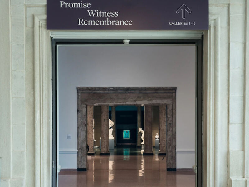 An entrance to a museum gallery with marble statues and a painting of a woman in the distance