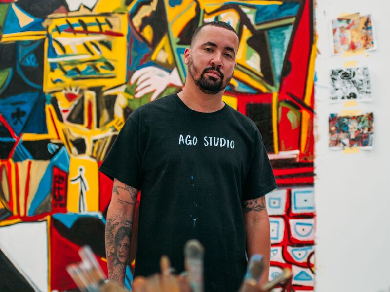 An artist stands in front of a colorful work of art in his studio