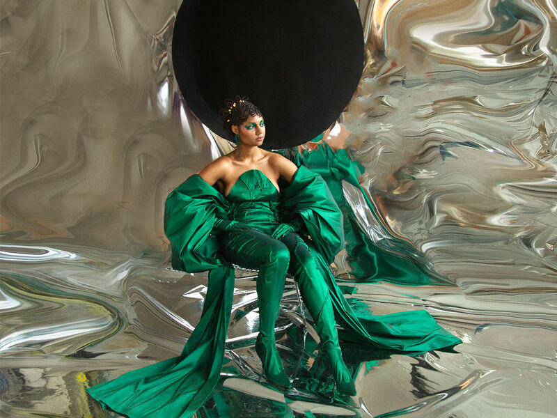 Person in an ornate green outfit sitting amongst a reflective backdrop