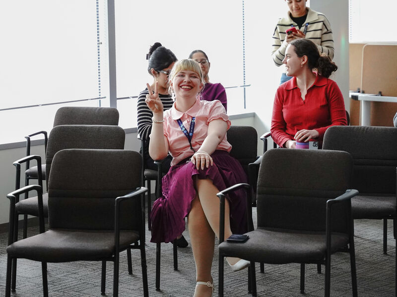 Lauren Nickels sits in a meeting room smiling and holding up a peace sign, surrounded by coworkers