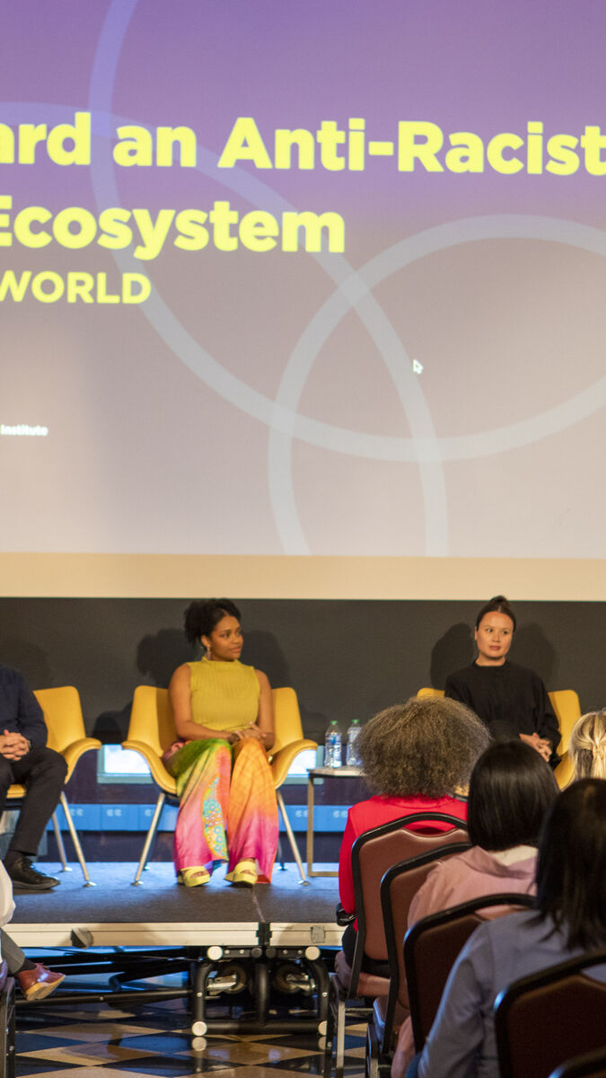 Screen reads “Toward an Anti-Racist Art Ecosystem in the World” with a panel of presenters in front and students in the audience.