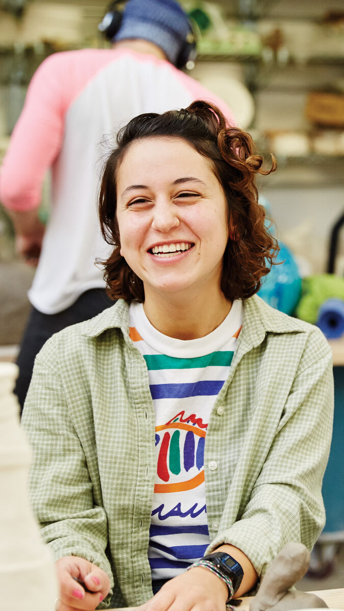 A person smiling and sitting at a throwing wheel in a ceramics studio. 