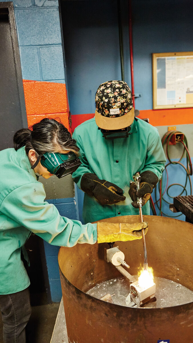 Two people wearing green protective smocks while heating up metal in a furnace. 