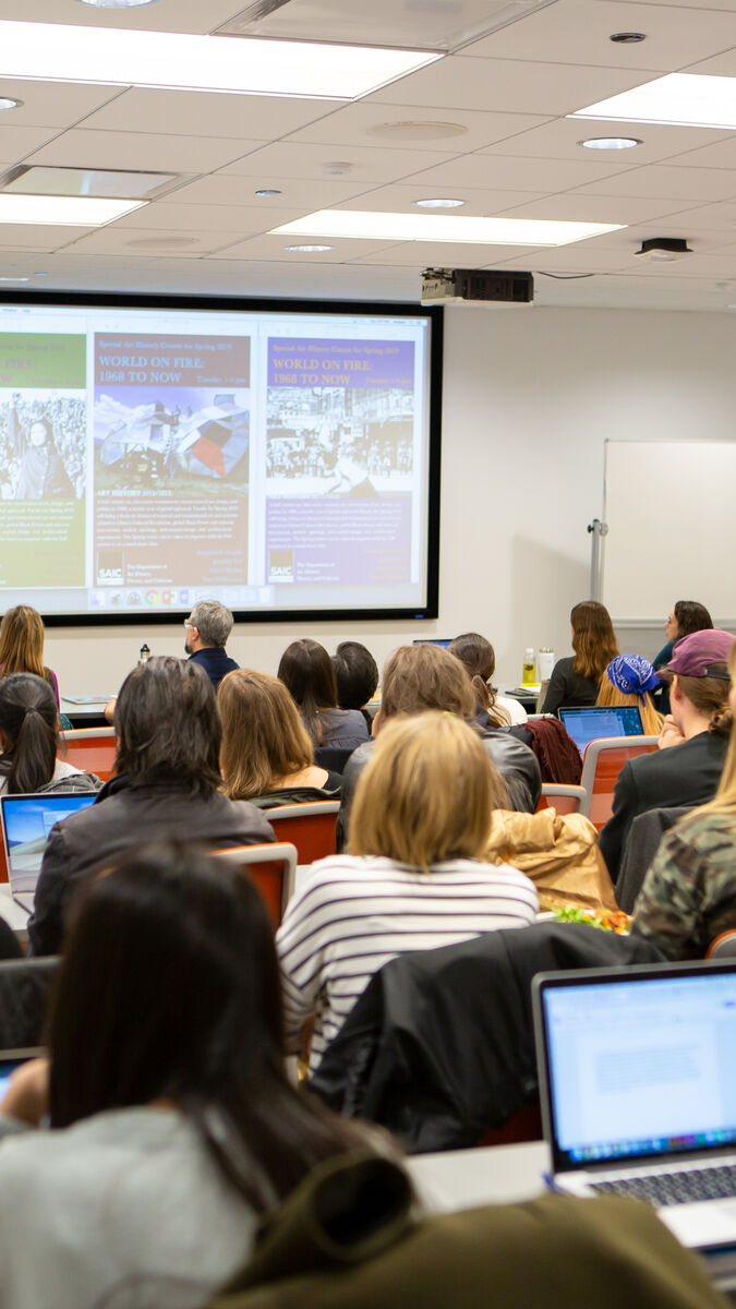 A classroom full of students facing a professor and projector.\