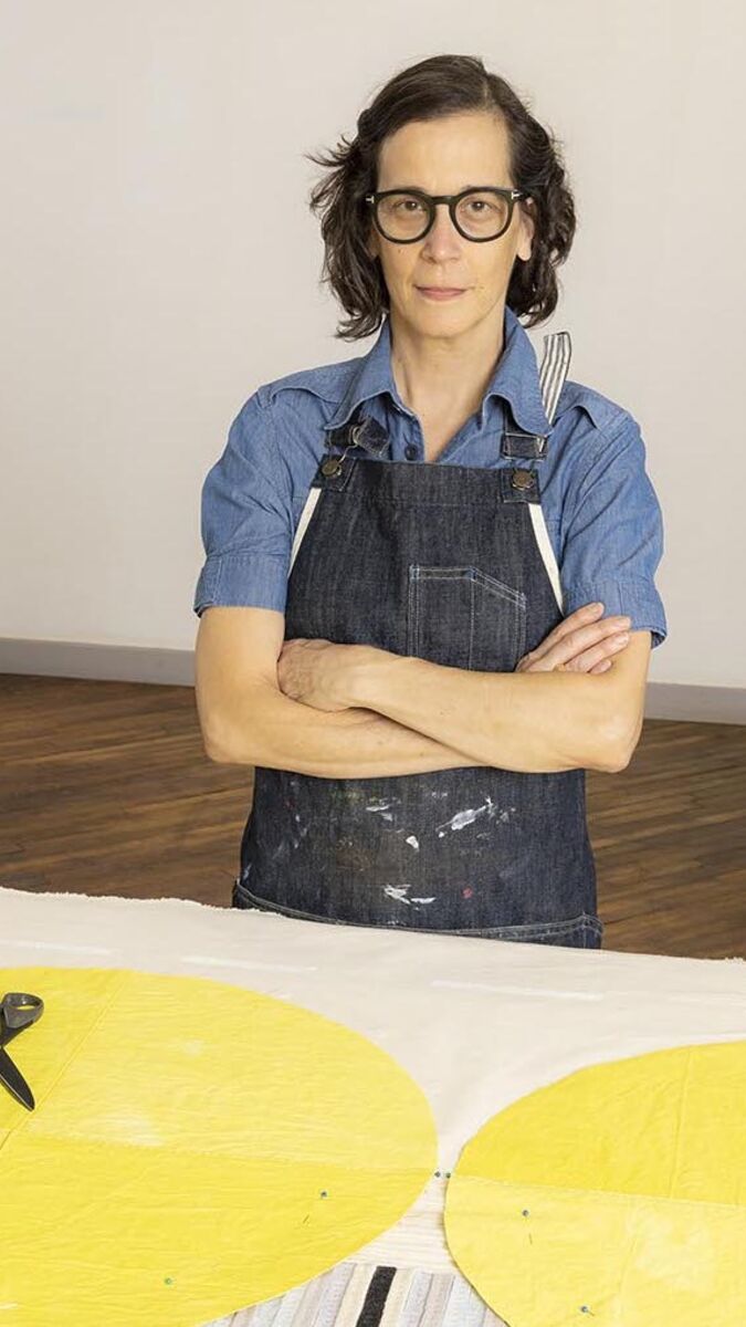 Diana stands, arms crossed, wearing a short sleeve denim shirt beneath a dark denim apron.