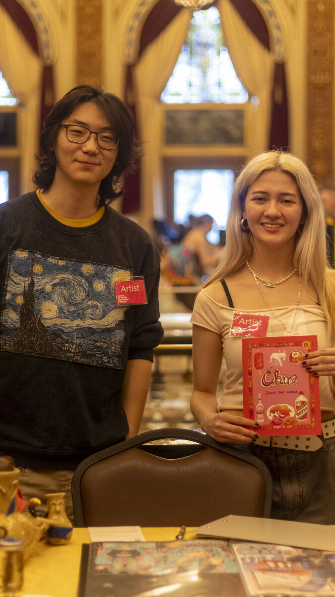 Two students at the Art Sale wearing "Artist" badges smiling and standing behind a table of various works of art. 