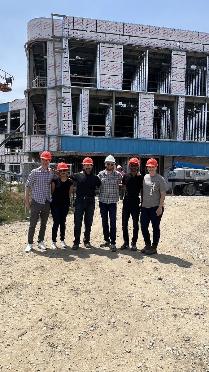 Six adults of varying ethnicities and genders wear hard hats and stand in front of a construction site.