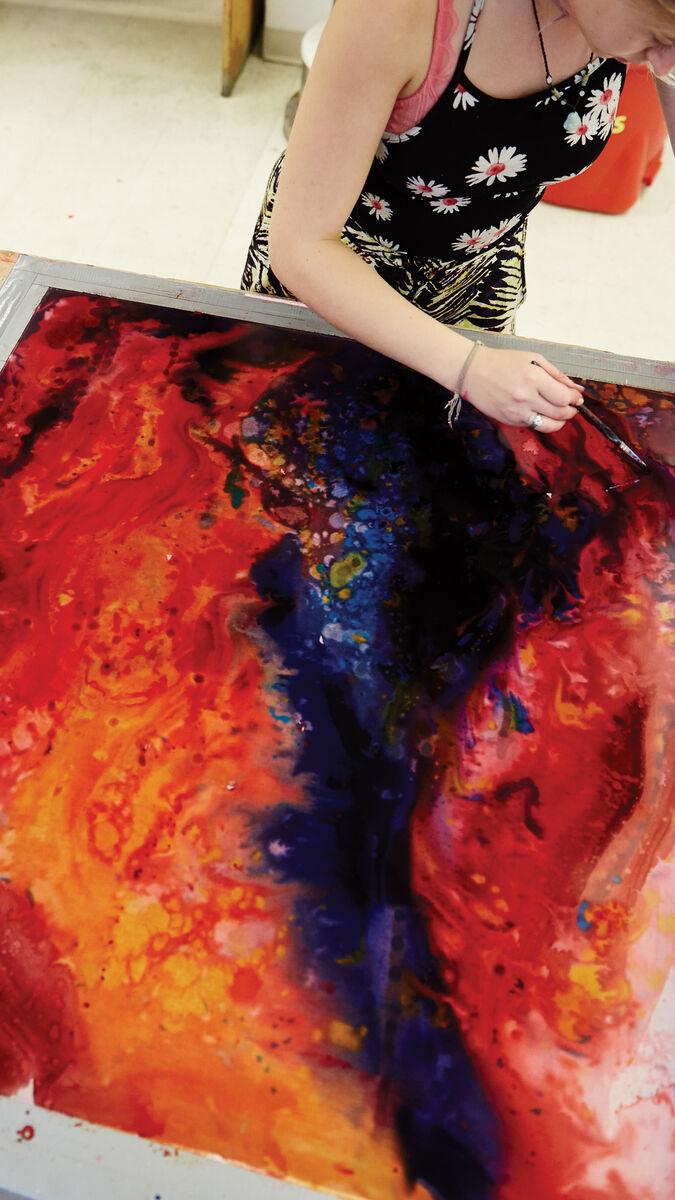 A person working on a colorful painting with paints nearby.