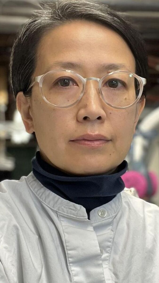 Mie Kongo faces the camera while crossing her arms. She is wearing a gray shirt and glasses with clear frames. Her studio can be seen in the background.