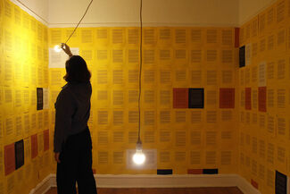 A person standing in a room illuminated by two lightbulbs with the walls covered in a grid of yellow, black, white, and red paper with text. 