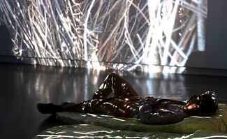A performer laying on a mat in a gallery space, with abstracted shapes projected onto them and the wall in the background
