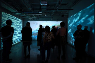 Numerous people in a dark room viewing an art installation of bluescale projections on three walls