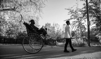 Black and white photograph by Cecil McDonald, Jr. of two Black men. One is pulling a carriage that the other sits in. 