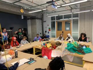 Students sit in tables arranged in a circle in a classroom, working on art projects.