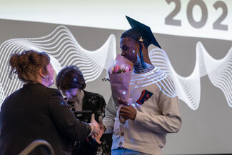 A student wearing a graduation cap and holding flowers shakes hands with an administrator.