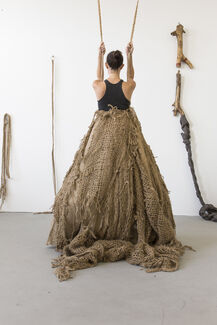 A person wearing a woven and sculpted skirt holds two ropes suspended from the ceiling. 