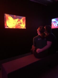 Two people view colorful light projections by Valerie Xanos in a dark gallery space. 