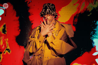 Bad Bunny posing with an embellished hat and nails in front of a red, black, orange, and blue backdrop. 