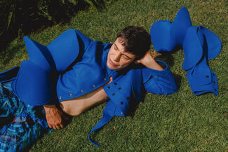 An adult person wearing bright blue designs by Edgar Aguilera laying in the grass surrounded by greenery. 