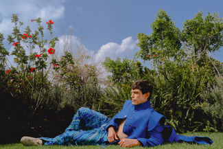 An adult person wearing bright blue designs by Edgar Aguilera laying in the grass surrounded by greenery. 