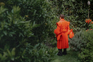 An adult person wearing bright orange designs by Edgar Aguilera standing and surrounded by greenery. 