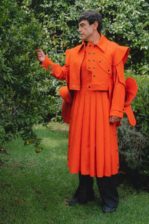 An adult person wearing bright orange designs by Edgar Aguilera standing and surrounded by greenery. 