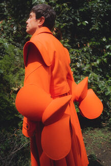 An adult person wearing bright orange designs by Edgar Aguilera standing and surrounded by greenery. 