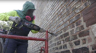 A person wearing a mask and protective clothing drills into a brick wall. 