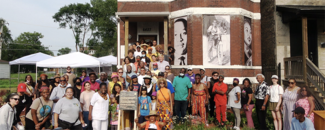 Dozens of people varying in age, ethnicity and gender stand outdoors in front of a home featuring black and white portraits of Black historical figures. 