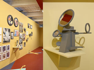 Gallery installation by Amy Yoes featuring geometric shapes in yellow, blue, and red; clocks; and mirrors. 