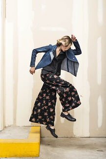 An adult person with a fair skin tone and blonde/brown hair models a denim jacket, black button-up and floral pants while jumping off a curb. 