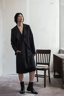 An adult person with pulled back dark hair and a fair skin tone, models a black blazer, black midi dress and black combat boots. 